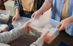 Obraz przedstawia osobę podającą przezroczyste plastikowe pudełko z produktami żywnościowymi innej osobie