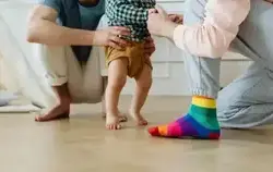 Na zdjęciu dwójka dorosłych ludzi oraz dziecko. Dorośli w przykucniętej pozycji, dziecko w trakcie chodzenia od mężczyzny do kobiety