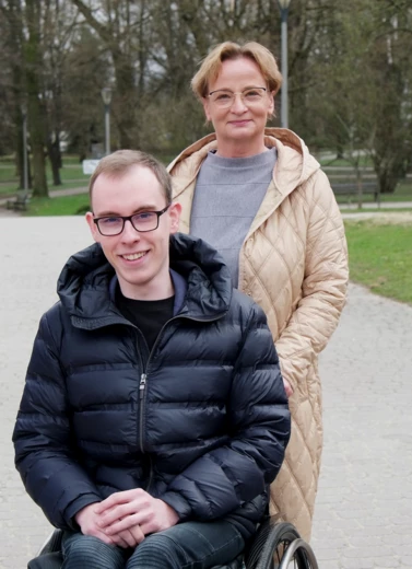 Zdjęcie przedstawia młodego mężczyznę na wózku inwalidzkim, jest uśmiechnięty, za nim stoi kobieta w krótkimi blond włosami