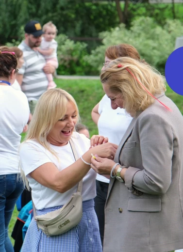 Zdjęcie przedstawia dwie uśmiechnięte kobiety w letnim otoczeniu, na tle bawiących się osób