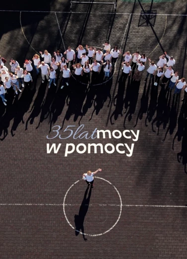 Zdjęcie przedstawia dużą grupę ludzi w ujęciu z powietrza - drona. Stoją na betonowym boisku, na środku widnieje napis "35 lat mocy w pomocy"