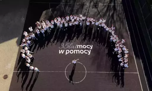 Zdjęcie przedstawia grupę osób stojących na betonowym boisku sportowym, na środku widnieje napis '35 lat mocy w pomocy'