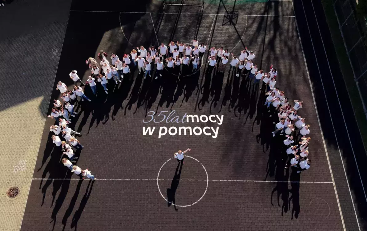 Zdjęcie przedstawia grupę osób stojących na betonowym boisku sportowym, na środku widnieje napis '35 lat mocy w pomocy'
