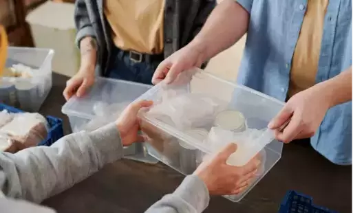 Obraz przedstawia osobę podającą przezroczyste plastikowe pudełko z produktami żywnościowymi innej osobie