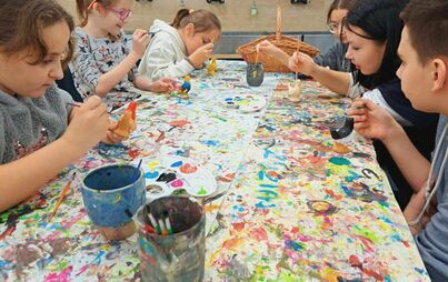 Na zdjęciu grupa dzieci siedzących przy stole, w rękach mają pędzle, malują